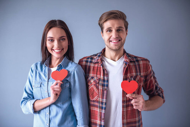 Счастливая молодая пара держит красные бумажные сердечки, смотрит в камеру и улыбается, на сером фоне
 - Фото, изображение