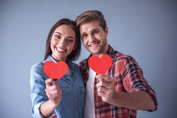 Happy young couple is holding red paper hearts, looking at camera and smiling, on gray background - Photo, Image