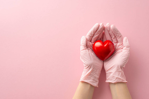 Temat medyczny: Pierwsza osoba nad głową ujęcie rąk młodej kobiety ubranych w rękawiczki chirurgiczne, kołyszące czerwone serce, symbolizujące macierzyństwo, na pastelowym różowym tle z miejscem na tekst lub reklamy - Zdjęcie, obraz