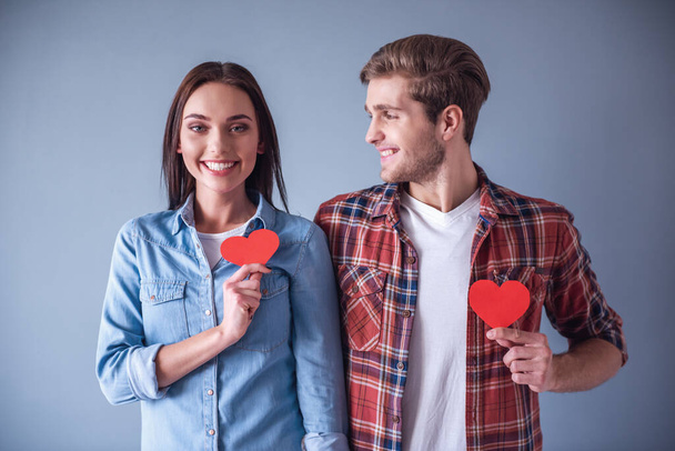 Счастливая молодая пара держит красные бумажные сердца и улыбается, на сером фоне
 - Фото, изображение