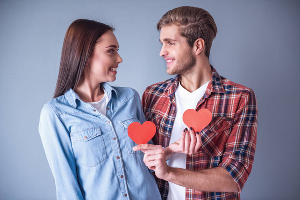 Happy young couple is holding red paper hearts, looking at each other and smiling, on gray background - Photo, Image