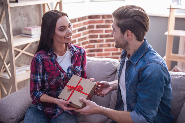Bello giovane uomo sta dando un regalo alla sua bella ragazza mentre sono seduti sul divano a casa
 - Foto, immagini