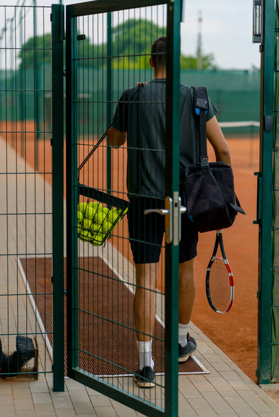 Genç profesyonel oyuncu koçu raket ve basket tenis toplarıyla antrenmana başlamadan önce açık hava tenis kortunda duruyor. - Fotoğraf, Görsel