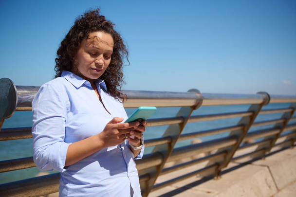 Pewna siebie bizneswoman w średnim wieku, korzystająca z telefonu komórkowego, sprawdzająca aplikację mobilną, stojąca na tle morza. Ludzie. Interesy. Komunikacja online - Zdjęcie, obraz