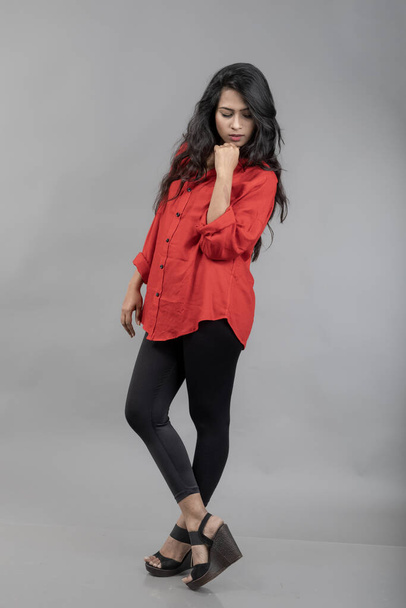 Modello giovane donna indiana in abbigliamento casual su sfondo grigio - foto stock. Modello lungo dai capelli neri con camicia rossa e jeans neri. - Foto, immagini