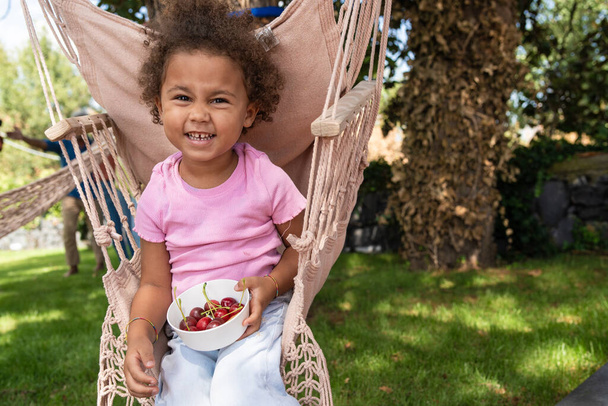 Egy vidám hároméves lány göndör hajjal ül egy kötélhintaszékben a kertben, kezében egy tál cseresznyével. Kaukázusi és afrikai származású, melegen néz a kamerába egy napos nyári napon.. - Fotó, kép