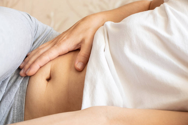 A girl lies on the bed with sore stomach and holds her stomach, acute pain in the abdomen, sick - Photo, Image