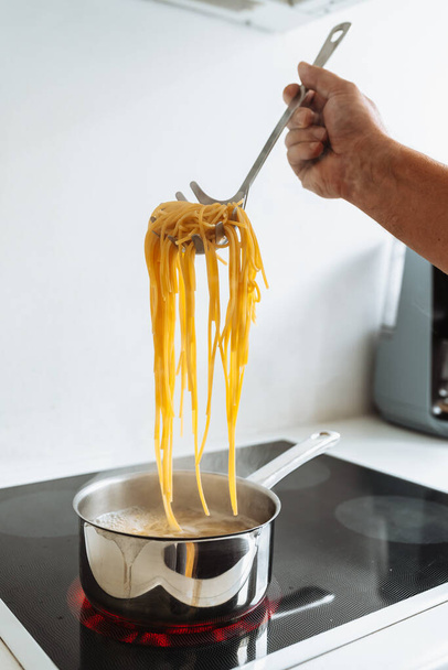 Ο άνθρωπος μαγειρεύει μακαρόνια στο σπίτι στην κουζίνα. μαγειρεύει με καφέ ποδιά, δοκιμάζει έτοιμα μακαρόνια, κρατάει σπαγγέτι σε ειδικό κουτάλι πάνω από βραστό νερό. Ιταλική κουζίνα στο σπίτι έννοια - Φωτογραφία, εικόνα