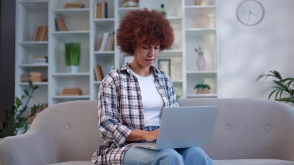 Mooie Afro-Amerikaanse vrouw met krullend haar typen op draadloze laptop en glimlachen terwijl zitten op een grijze bank. Mooie vrouwelijke freelancer in casual kleding genieten van afgelegen werk thuis. - Video