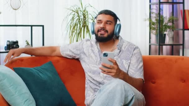 自宅でソファーでダンスするワイヤレスヘッドフォンで幸せなリラックスしたインディアン男性は,スマートフォンの娯楽で好きなエネルギッシュディスコロックNロールミュージックを聞くことを選択します. 人々 週末の活動 - 映像、動画