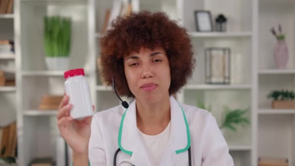 Afrikkalainen amerikkalainen nainen langattomissa kuulokkeissa osoittaa valkoista purkkia pillereitä puhuessaan potilaan kanssa verkossa lääketieteellisessä kaapissa. Naisterapeutti antaa lääkesuosituksensa videovuokrauksen aikana. - Materiaali, video