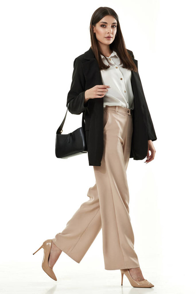 attractive lady holding bag walking in black blazer and beige pants, high-heels isolated on white background. Full-length - 写真・画像