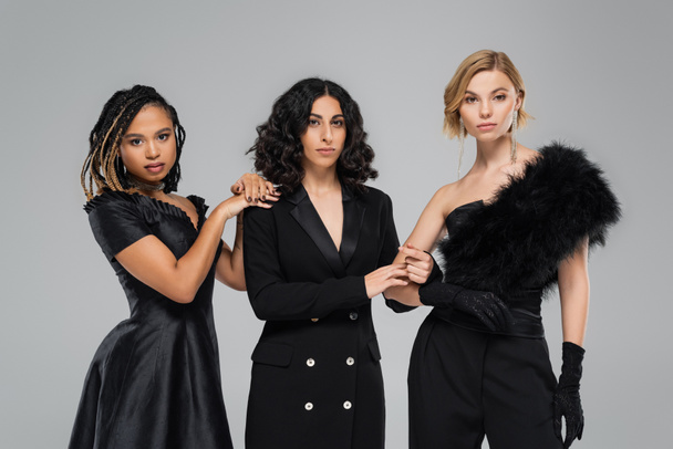 drei stylische Frauen in ganz schwarzer eleganter Kleidung auf grau, Gruppe moderner multirassischer Freundinnen - Foto, Bild
