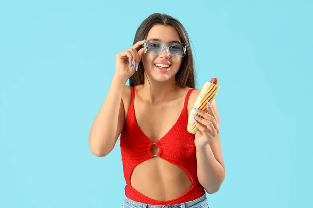 Hübsche junge Frau in Bademode und mit leckerem französischen Hot Dog auf blauem Hintergrund - Foto, Bild