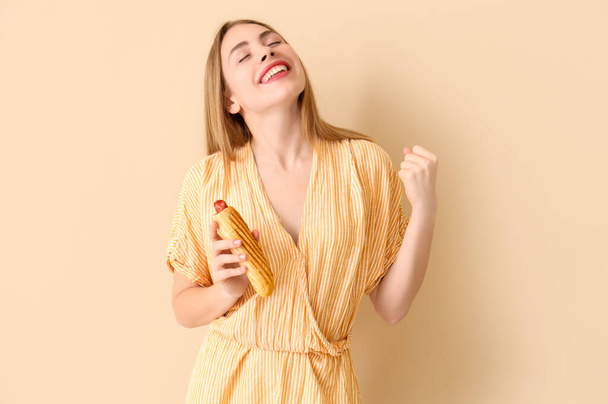 Ευτυχισμένη νεαρή γυναίκα με νόστιμο γαλλικό χοτ ντογκ σε μπεζ φόντο - Φωτογραφία, εικόνα