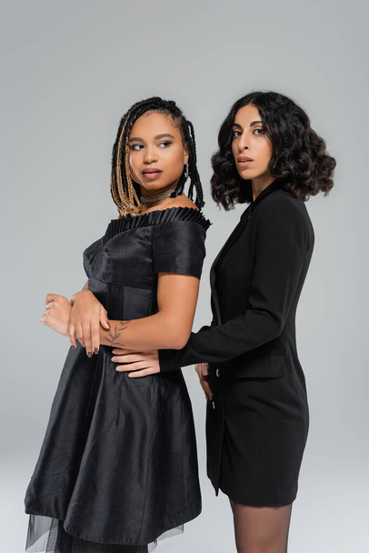 разнообразие и мода, две многорасовые девушки в черной элегантной одежде позируют на сером фоне - Фото, изображение