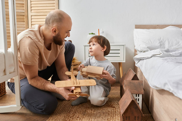 веселый современный отец и сын в уютной домашней одежде играют с деревянными игрушками и смотрят друг на друга - Фото, изображение