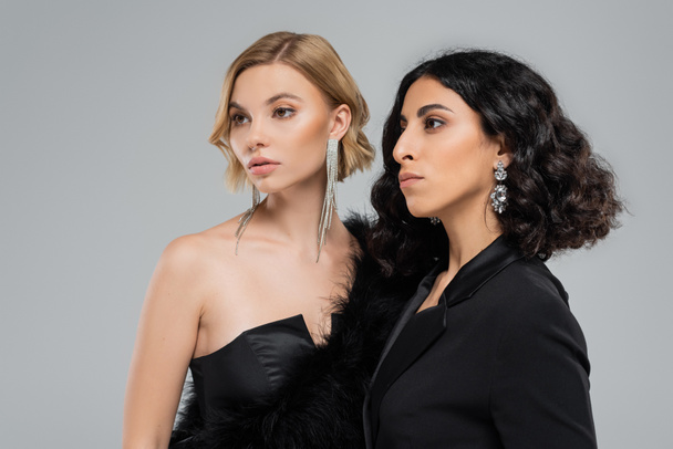 portrait of multiracial girlfriends in black elegant attire and silver earrings on grey backdrop - Photo, Image