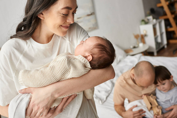 focus op gelukkig moeder kijken naar haar pasgeboren met wazig echtgenoot en kleine zoon spelen op de achtergrond - Foto, afbeelding