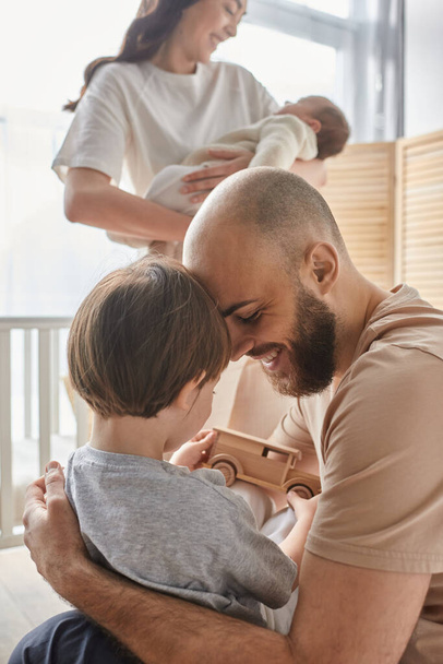 κάθετη εστιασμένη λήψη του πατέρα παίζει με το γιο του με θολή γυναίκα του με νεογέννητο στο φόντο - Φωτογραφία, εικόνα