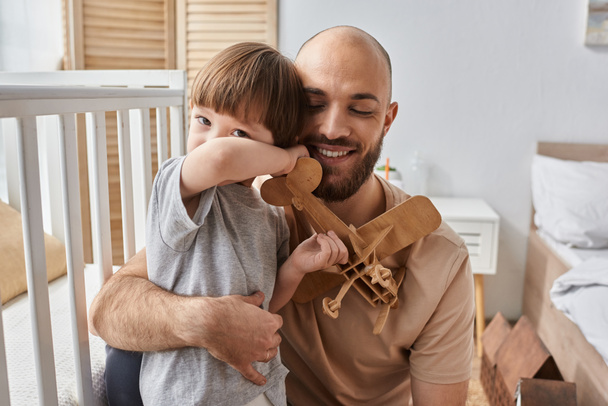 glücklicher gutaussehender Vater umarmt seinen kleinen Sohn herzlich beim Spielen mit Holzflugzeug, Familienkonzept - Foto, Bild