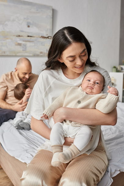 背中に彼女のぼやけた夫と小さな息子と彼女の新生児を抱えている母親の垂直ショット - 写真・画像