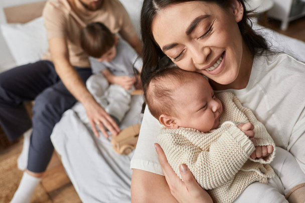 zaměřit se na veselou matku objímající svého novorozeného chlapečka vedle svého rozmazaného manžela a malého syna - Fotografie, Obrázek