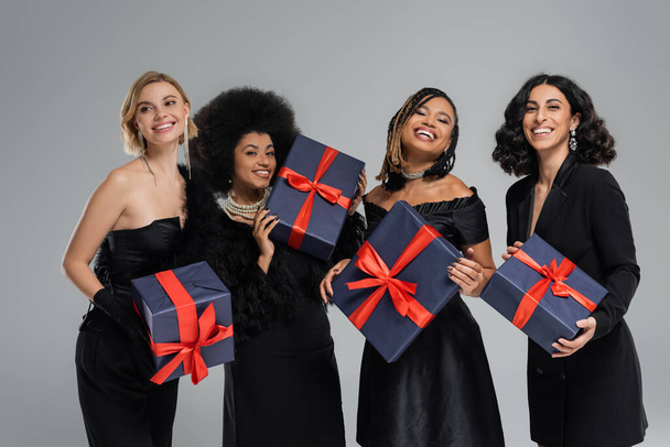 група веселих багатоетнічних жінок в чорному елегантному вбранні, що тримає святкові подарунки на сірому - Фото, зображення