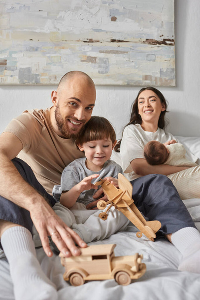 Fröhlicher bärtiger Vater umarmt seinen Sohn lächelnd in die Kamera mit seiner Frau und dem neugeborenen Baby vor der Kulisse - Foto, Bild