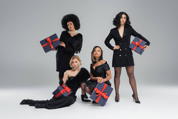 modelli multiculturali di moda in posa in nero elegante abbigliamento con scatole regalo di festa su grigio - Foto, immagini