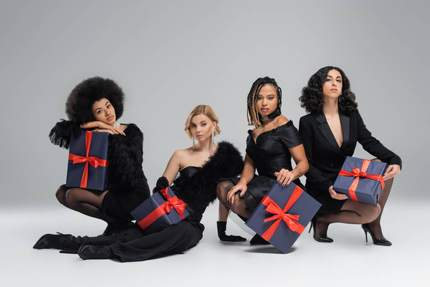 group of diverse multiracial girlfriends in total black stylish wear sitting with gift boxes on grey - Photo, Image