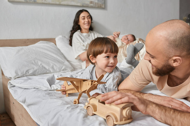 concentrarsi sul padre e piccolo figlio sdraiato sul letto con madre offuscata che tiene il neonato sullo sfondo - Foto, immagini