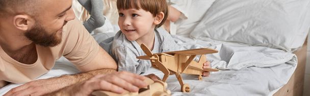vrolijke vader liggend op bed met zijn zoontje spelen houten speelgoed en lachen naar elkaar, spandoek - Foto, afbeelding