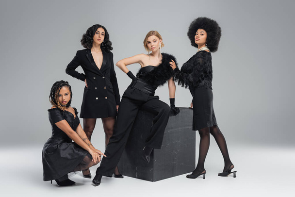 Charmante multikulturelle Damen in schwarzer eleganter Kleidung blicken in die Kamera in der Nähe von Dark Cube auf Grau - Foto, Bild