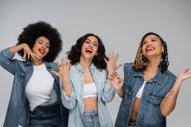 overjoyed multiracial girlfriends in blue denim clothes gesturing on grey, friendship and happiness - Photo, Image
