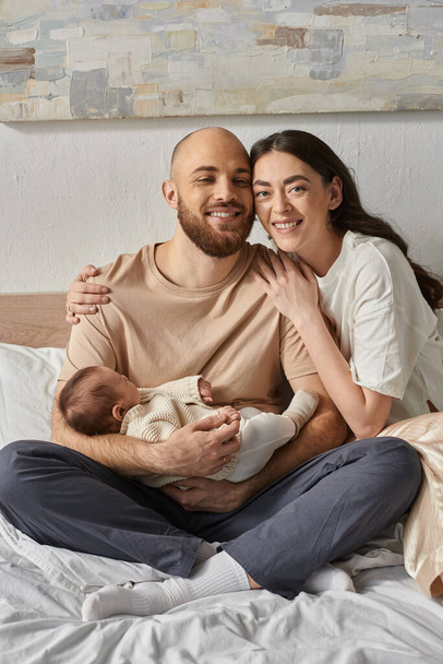 vertical shot of joyous parents sitting on bed smiling and holding their cute newborn baby, family - Photo, Image