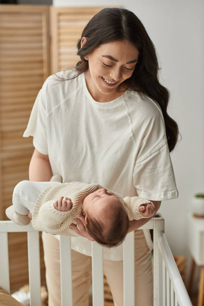 vertical shot of happy modern mother putting her cute newborn baby into his crib, family concept - Fotó, kép