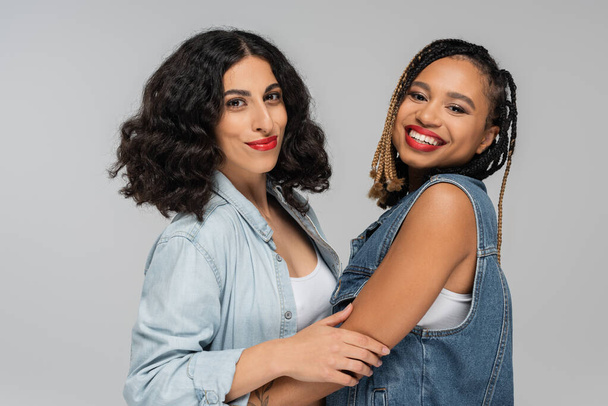 две беззаботные многорасовые подружки в стильной джинсовой одежде, улыбающиеся в камеру на сером, вместе - Фото, изображение