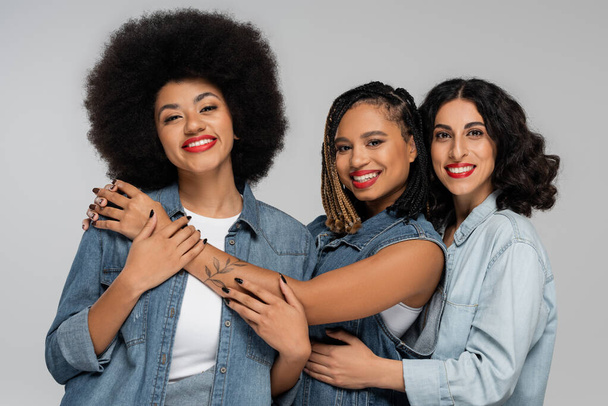 stylish and smiley multiethnic girlfriends in blue jeans wear looking at camera on grey, diversity - Photo, Image