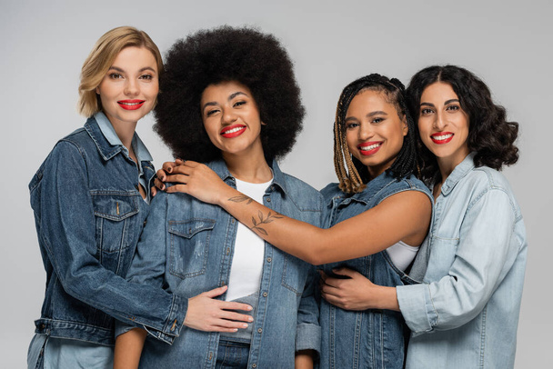 jolly and trendy multicultural girlfriends in blue denim wear smiling at camera on grey, friendship - Photo, Image