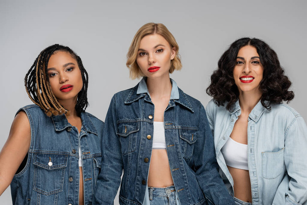 three attractive multiracial women in denim clothes looking at camera on grey, group portrait - Photo, Image