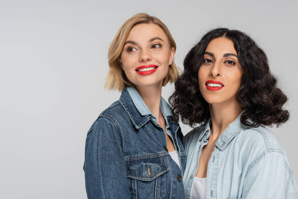 portrait of cheerful blonde and brunette multiracial girlfriends in blue jeans wear on grey backdrop - 写真・画像