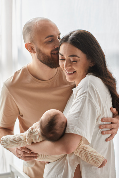 vertikale Aufnahme fröhlicher, glücklicher Eltern, die sich liebevoll umarmen und ihr neugeborenes Baby halten, Familie - Foto, Bild