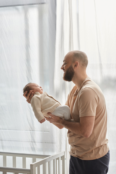 Vertikale Aufnahme eines glücklichen Vaters in kuscheliger Hauskleidung, der seinen neugeborenen Sohn hält und ihn liebevoll anlächelt - Foto, Bild