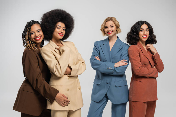 grupo de namoradas multiétnicas na moda em ternos multicoloridos sorrindo para a câmera em cinza, diversidade - Foto, Imagem