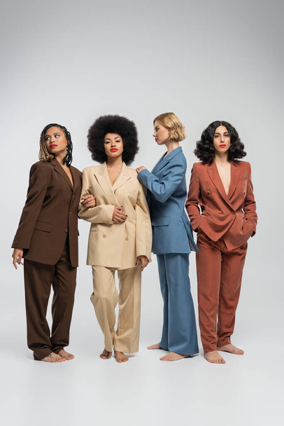 multiculturali donne scalze in abiti multicolori in piedi su sfondo grigio, fotografia di moda - Foto, immagini