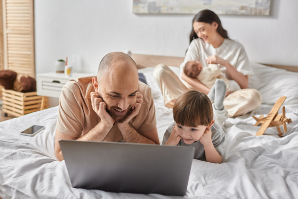 se concentrer sur le père heureux et le petit fils regarder un film sur ordinateur portable à côté de sa femme floue et bébé garçon - Photo, image