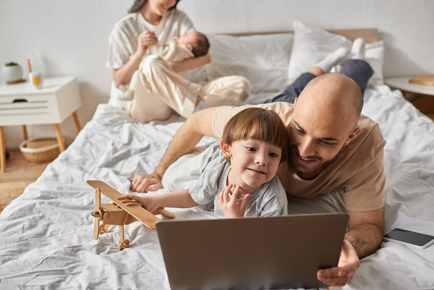 focus on little boy with his father looking at laptop next to his blurred mother and baby brother - Photo, Image