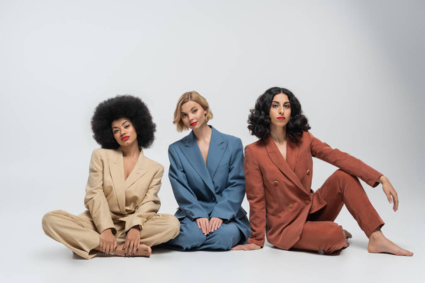Barfuß multiethnische Freundinnen in trendigen bunten Anzügen sitzen und blicken in die Kamera auf grau - Foto, Bild