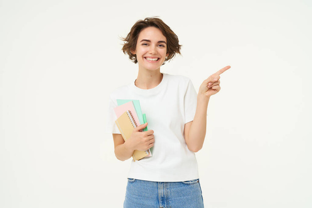 Portrait of brunette woman laughing, student with notebooks pointing at upper right corner, showing banner or advertisement, standing over white studio background. - Photo, Image